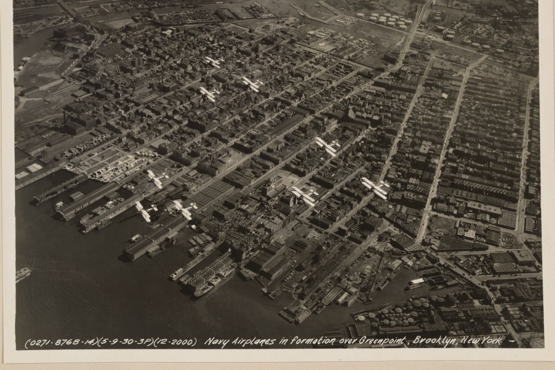 Black-and-white 1930 aerial photo of Navy biplanes flying over the Greenpoint waterfront's piers, rail yards, and tank farms.
