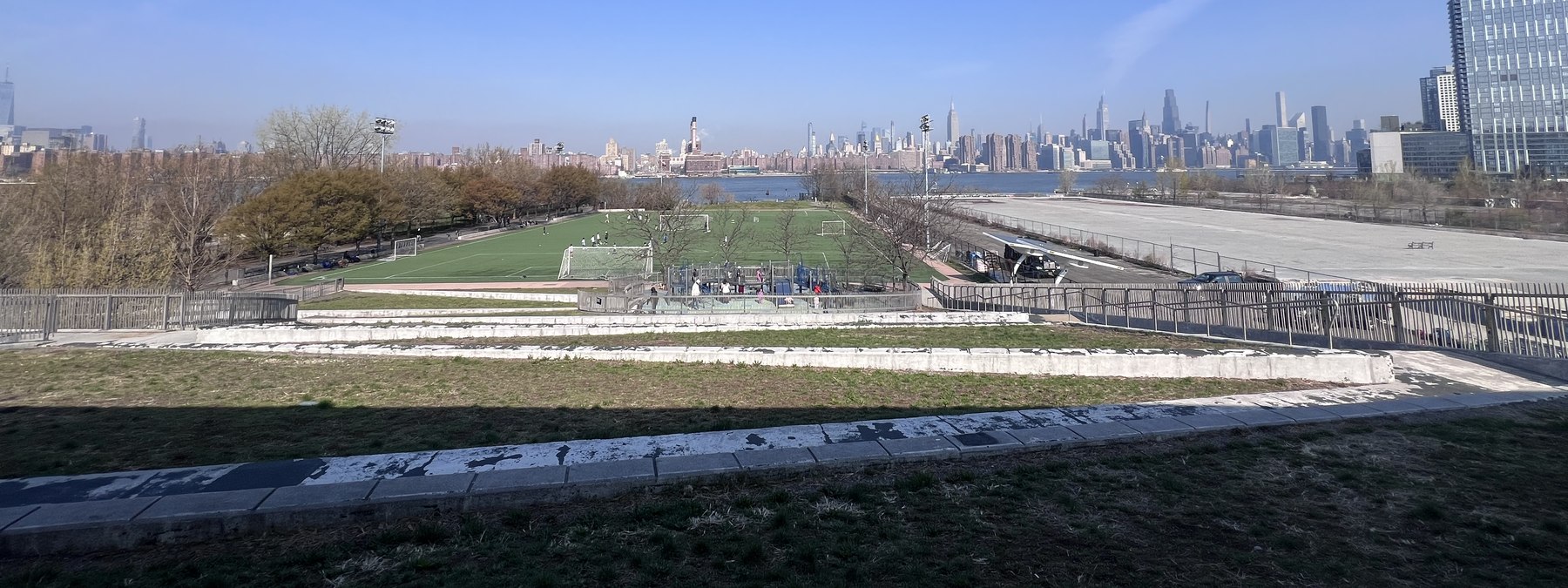 View from a green roof: a full soccer field on the left and a flat, empty asphalt lot on the right, with the East River and Manhattan skyline behind.