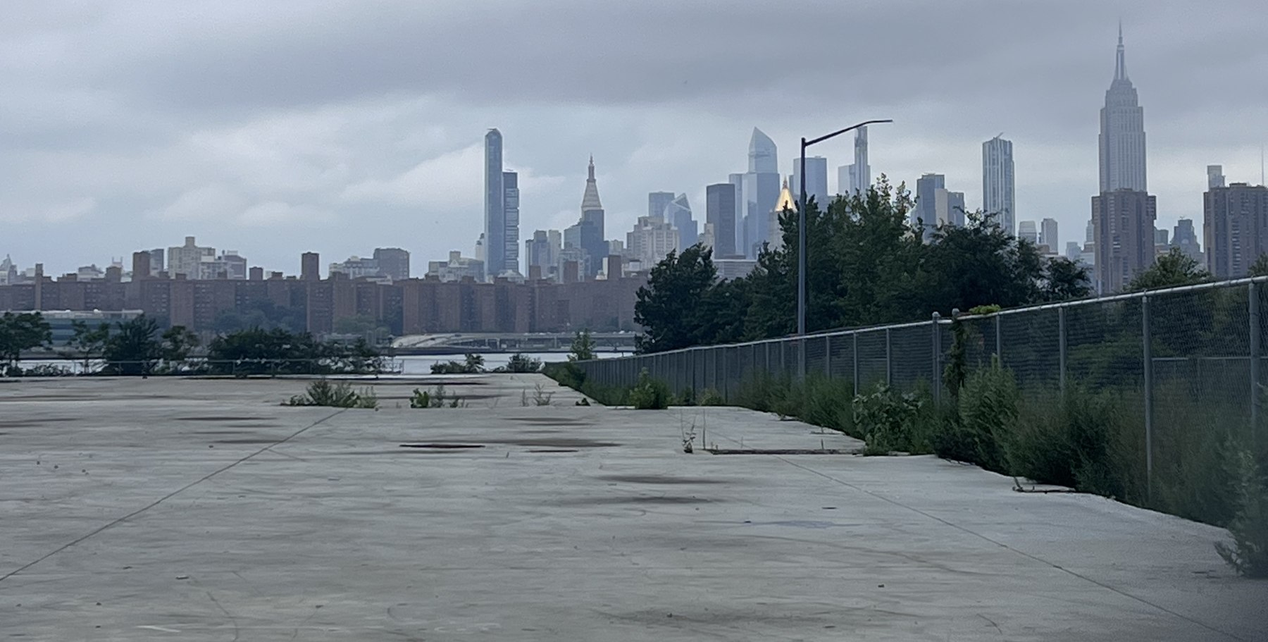 A chain-link fence around a vacant lot of cracked asphalt and weeds, with the Manhattan skyline visible across the river.